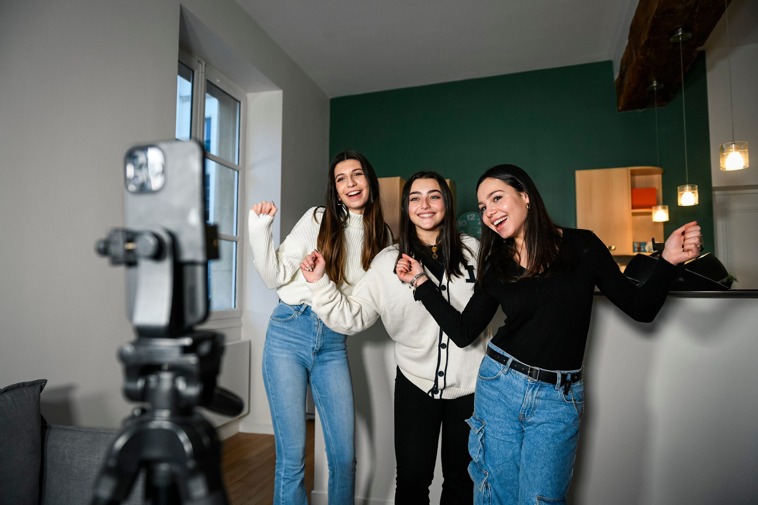 ParisFrance-June320233HappyWhite | クロスボーダーネクスト株式会社 Three young women pose for a selfie indoors, smiling at a camera on a tripod in a living room.