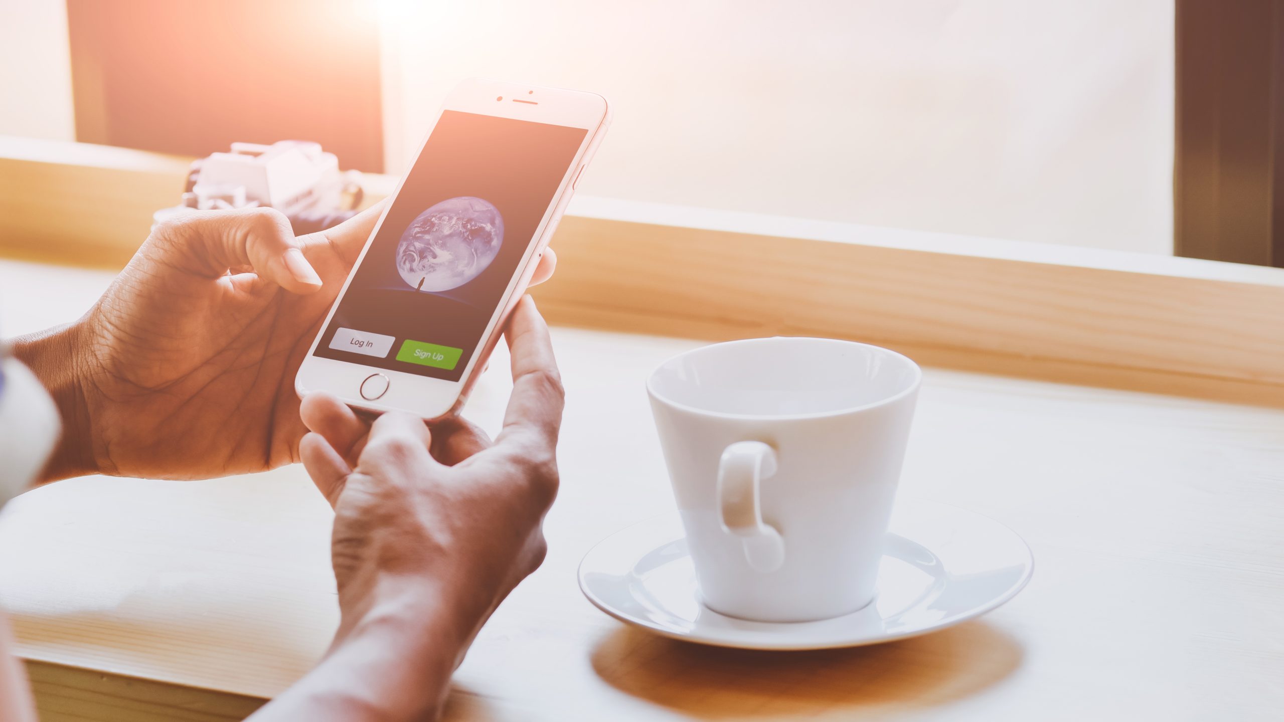 BangkokThailandFebruary32017YoungWomanHoldingIphone6s | クロスボーダーネクスト株式会社 Person holding a smartphone showing Earth on the screen, at a cafe with a white coffee cup nearby.