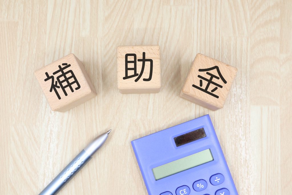 shutterstock_2488953327 1 | クロスボーダーネクスト株式会社 Three wooden blocks arranged on a light wood surface spell out the Chinese/Japanese term 補助金 (subsidy).