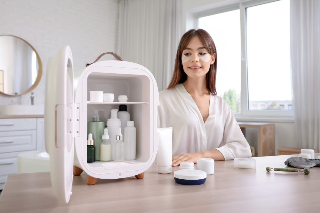 shutterstock_2578635175 1 | クロスボーダーネクスト株式会社 Woman with under-eye patches looking at an open mini skincare fridge filled with bottles and jars.