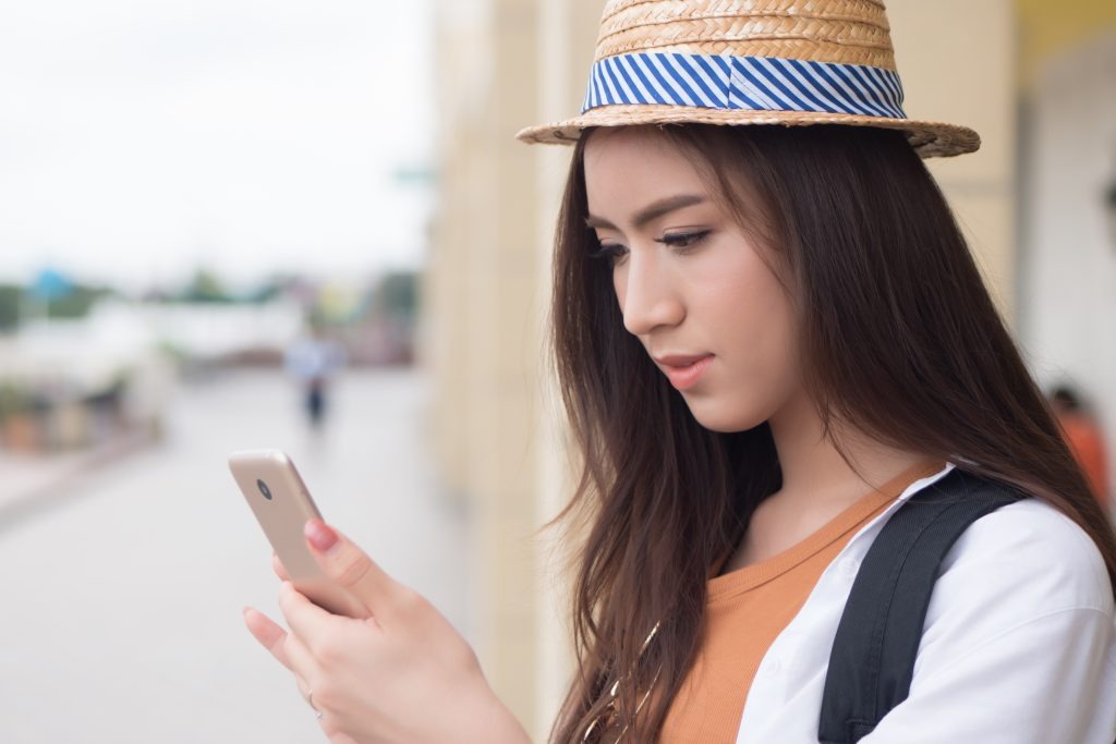 image | クロスボーダーネクスト株式会社 Woman wearing a straw hat and backpack, looking at her smartphone outdoors.
