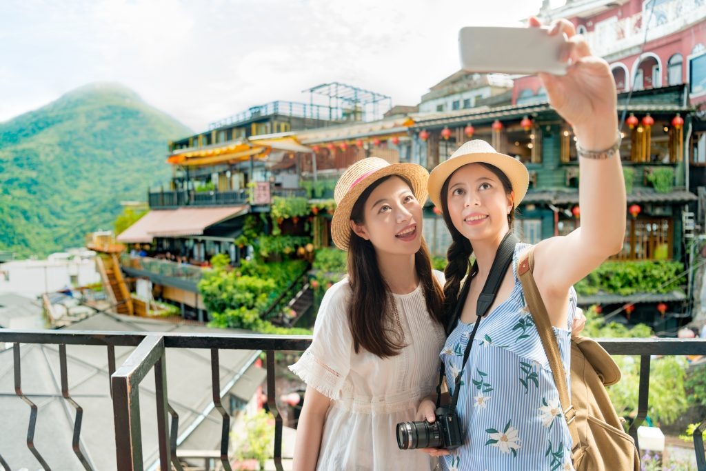 image | クロスボーダーネクスト株式会社 Two women wearing sun hats take a selfie on a balcony overlooking a colorful hillside town and green mountains behind them.