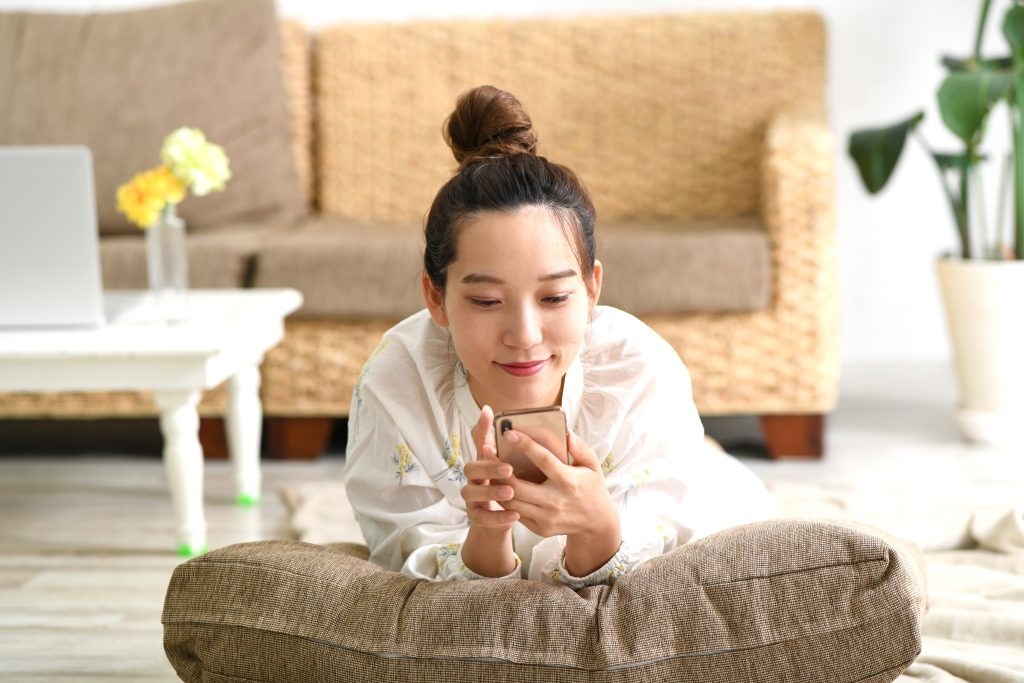 image | クロスボーダーネクスト株式会社 Woman lying on a floor cushion at home, looking at her smartphone with a laptop and plants in the background.