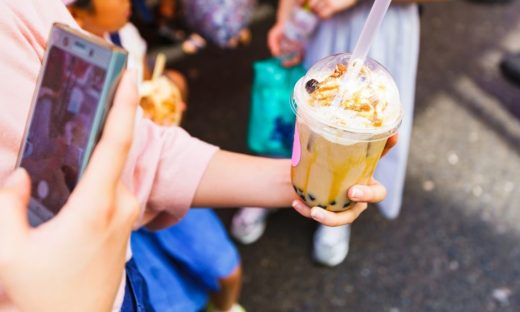image | クロスボーダーネクスト株式会社 A hand offers a bubble tea cup topped with whipped cream and caramel, while another person uses a smartphone to take a photo nearby to the scene.