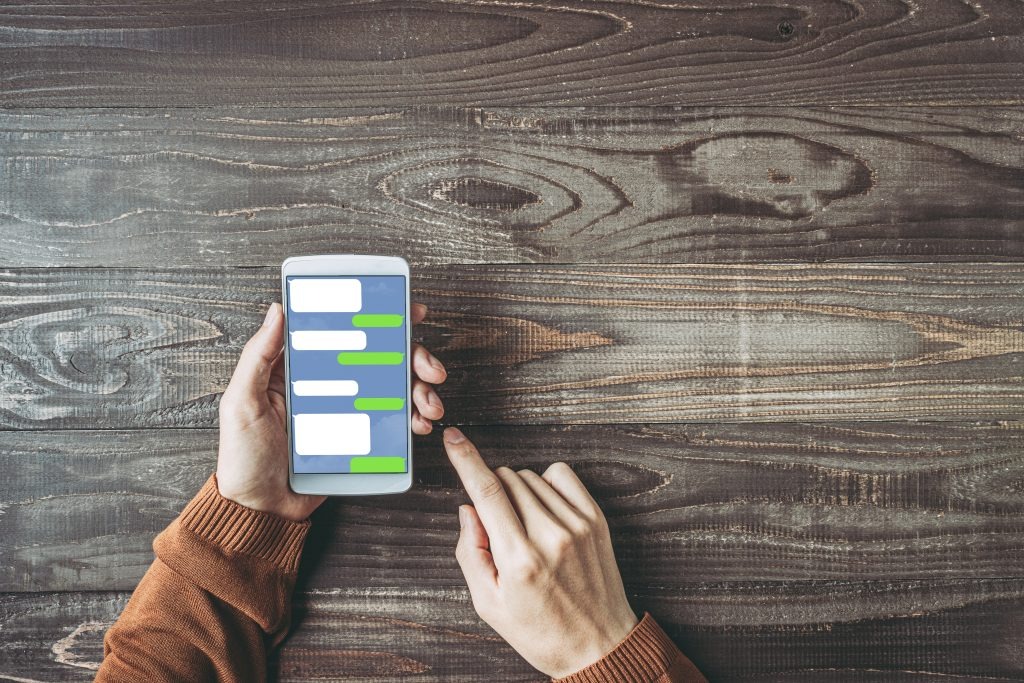 image | クロスボーダーネクスト株式会社 Person holding a smartphone with chat bubbles on screen while another hand points at it on a wooden table surface