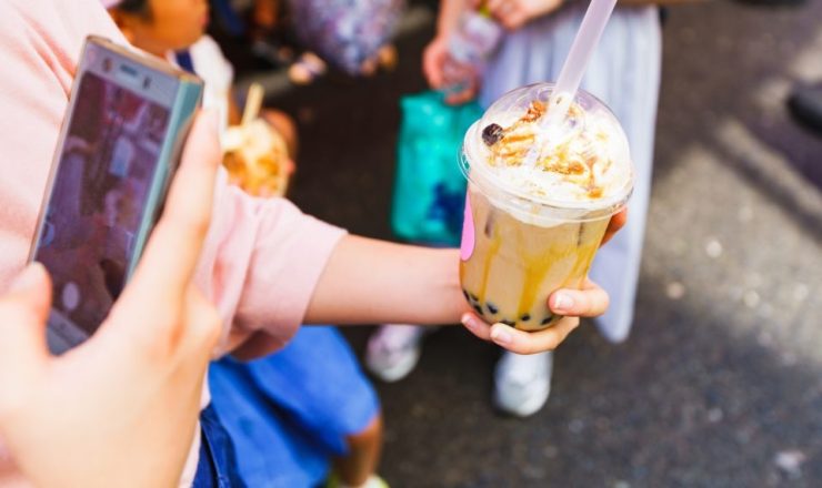A hand offers a bubble tea cup topped with whipped cream and caramel, while another person uses a smartphone to take a photo nearby to the scene.