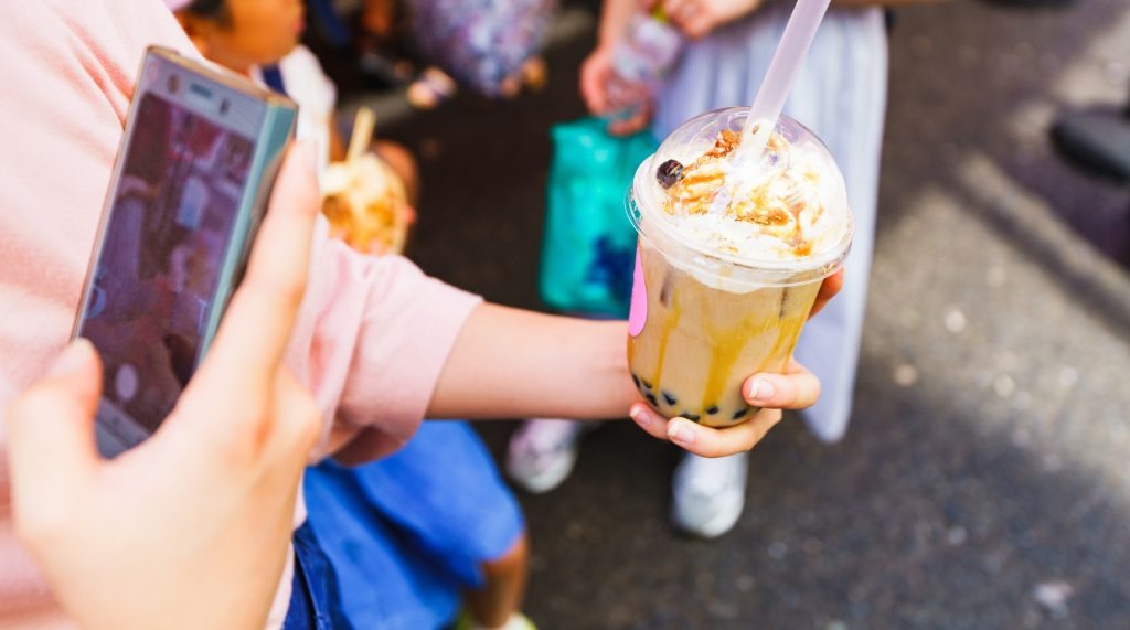 image | クロスボーダーネクスト株式会社 A hand offers a bubble tea cup topped with whipped cream and caramel, while another person uses a smartphone to take a photo nearby to the scene.