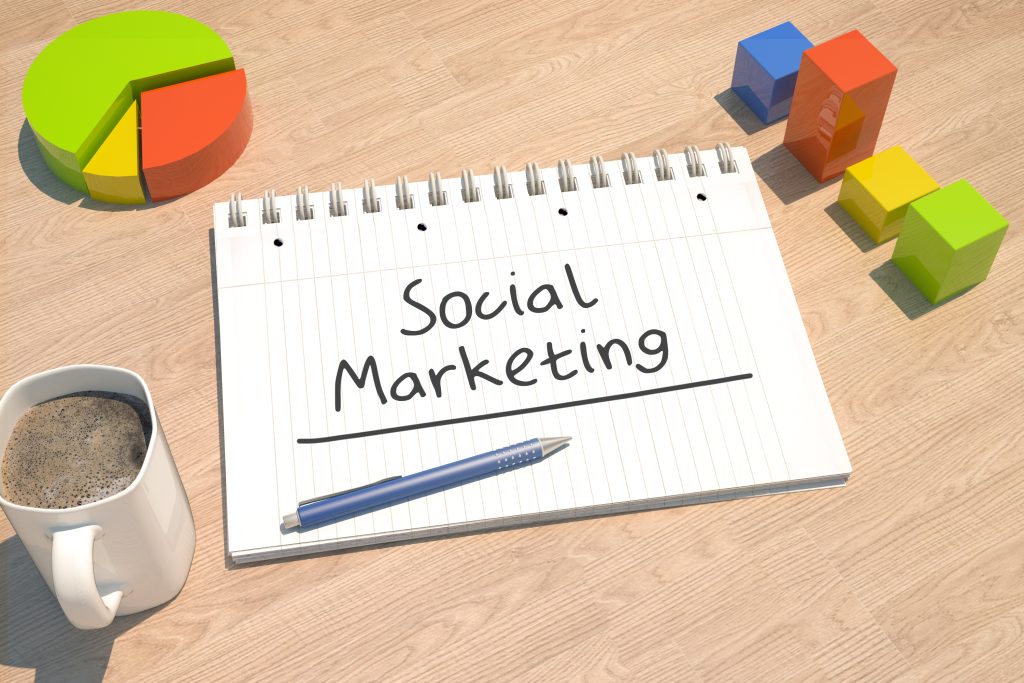 Notebook with 'Social Marketing' title, blue pen, and coffee cup on a wooden desk beside colorful 3D shapes and a pie chart