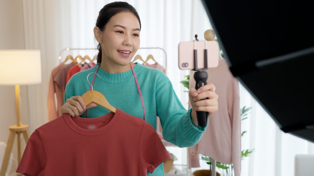 Woman filming a fashion haul with a smartphone on a tripod, wearing a teal sweater and holding a red shirt on a hanger.