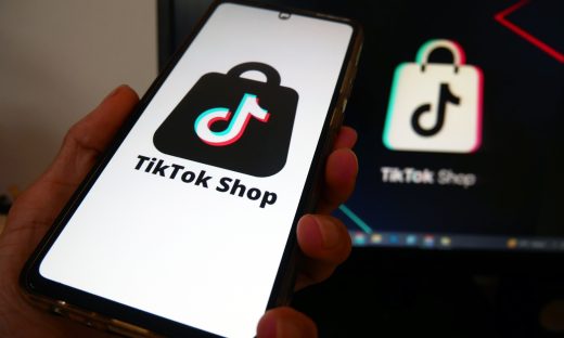 Hand holding a smartphone displaying the TikTok Shop logo on the screen, with a TikTok icon blurred in the background on a dark screen behind.
