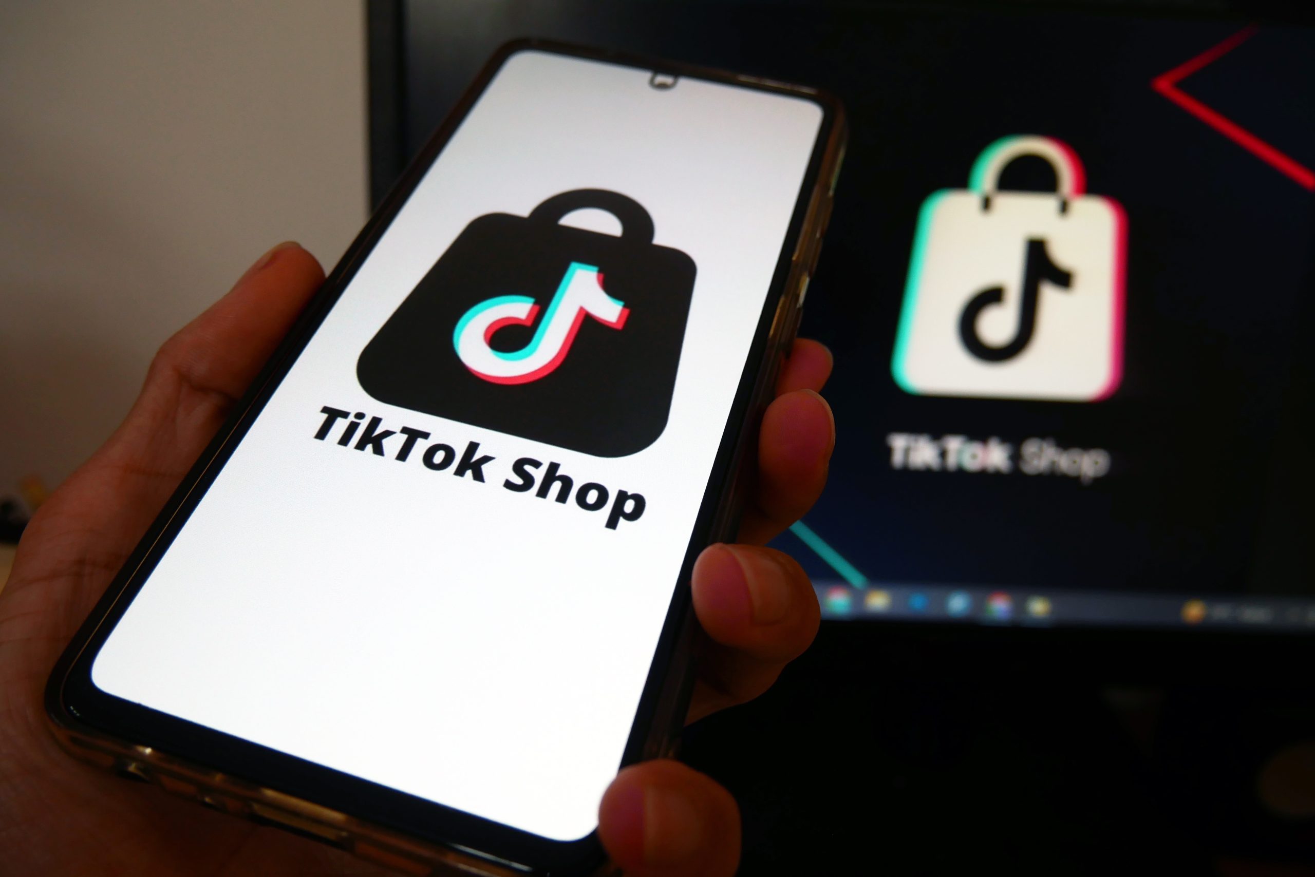 Hand holding a smartphone displaying the TikTok Shop logo on the screen, with a TikTok icon blurred in the background on a dark screen behind.