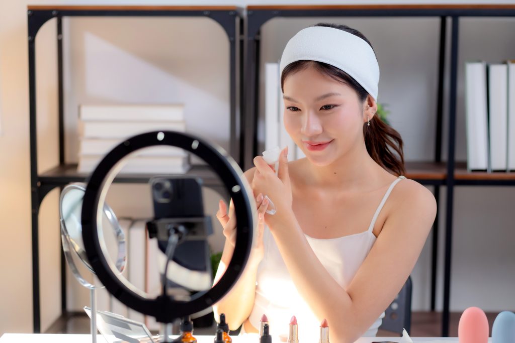 Woman applying lipstick in front of a ring light mirror, makeup products lined up on the table.