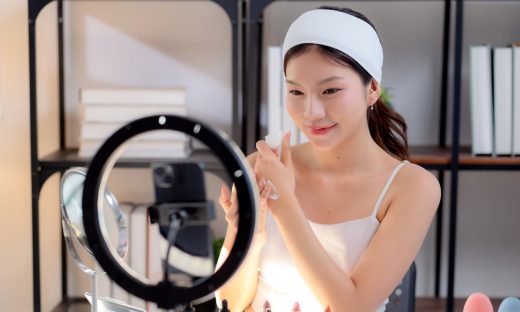 YoungAsianWomanPresentingAndReviewCosmeticProductWithOn | クロスボーダーネクスト株式会社 Woman applying lipstick in front of a ring light mirror, makeup products lined up on the table.