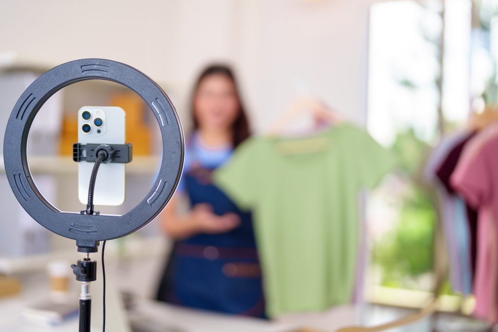 Smartphone mounted on a tripod inside a circular ring light, setup for video recording with a blurred person in the background