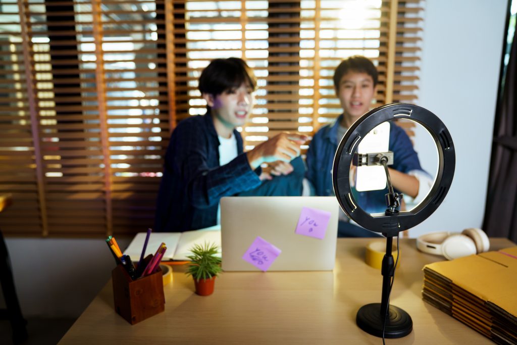 Two people sit at a desk with a laptop and sticky notes, ring light in the foreground suggesting a recording or livestream setup.