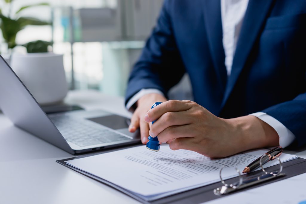Business professional stamping documents at a laptop desk in an office.