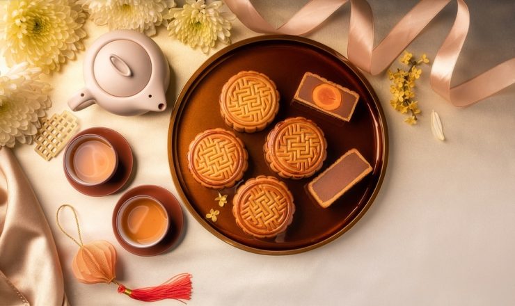 Round brown platter of mooncakes with two pieces cut open, surrounded by tea cups and a teapot on a satin tablecloth.