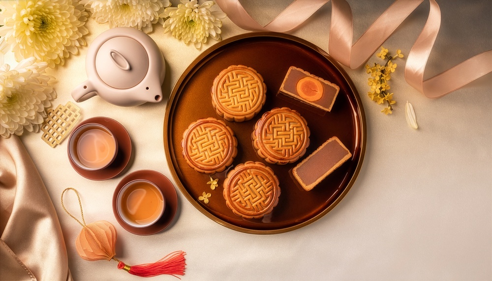 MinimalMooncakeFlatLayWithTeaAndLanternTopView | クロスボーダーネクスト株式会社 Round brown platter of mooncakes with two pieces cut open, surrounded by tea cups and a teapot on a satin tablecloth.